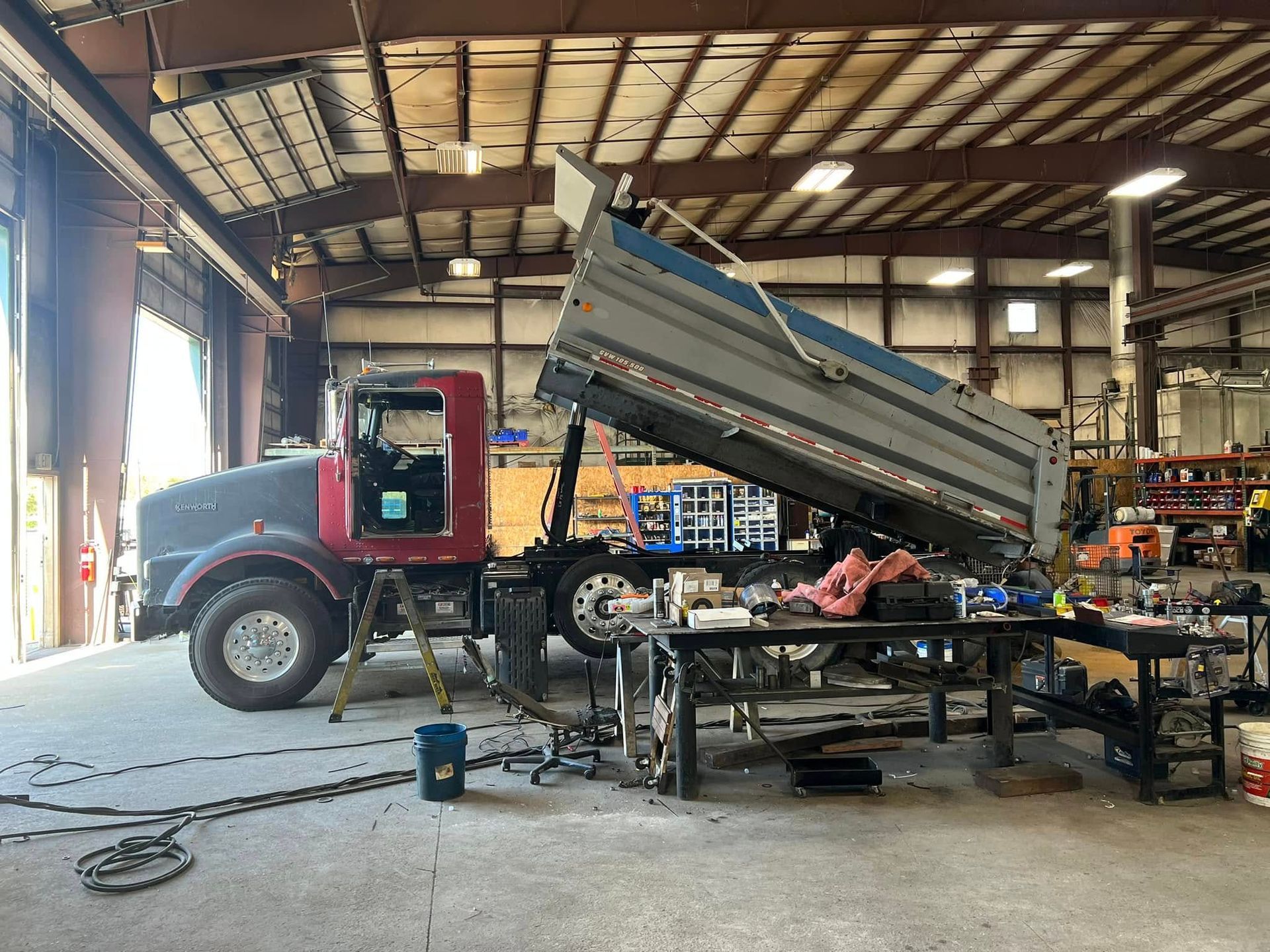 Red dump truck with raised bed inside a repair shop, surrounded by tools and equipment.