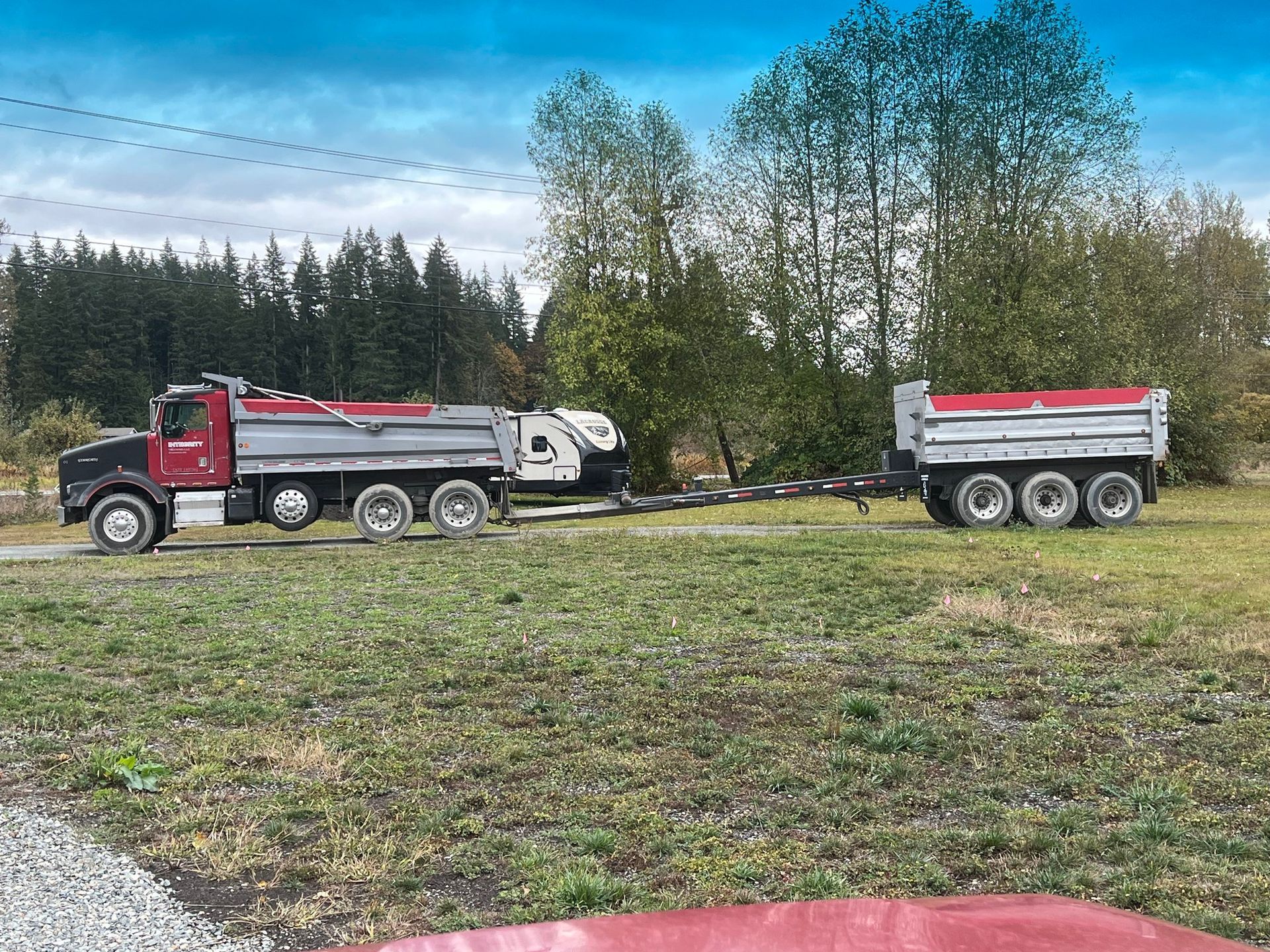 Two dump trucks parked in a grassy field with trees and a cloudy sky in the background.