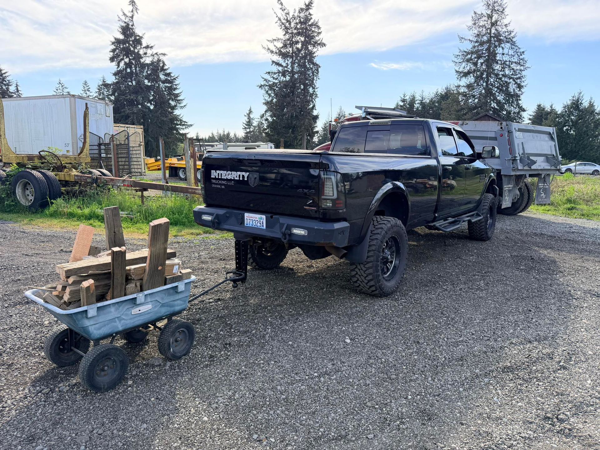 Black lifted pickup truck parked on gravel beside a small blue wagon with wood planks and trees in the background