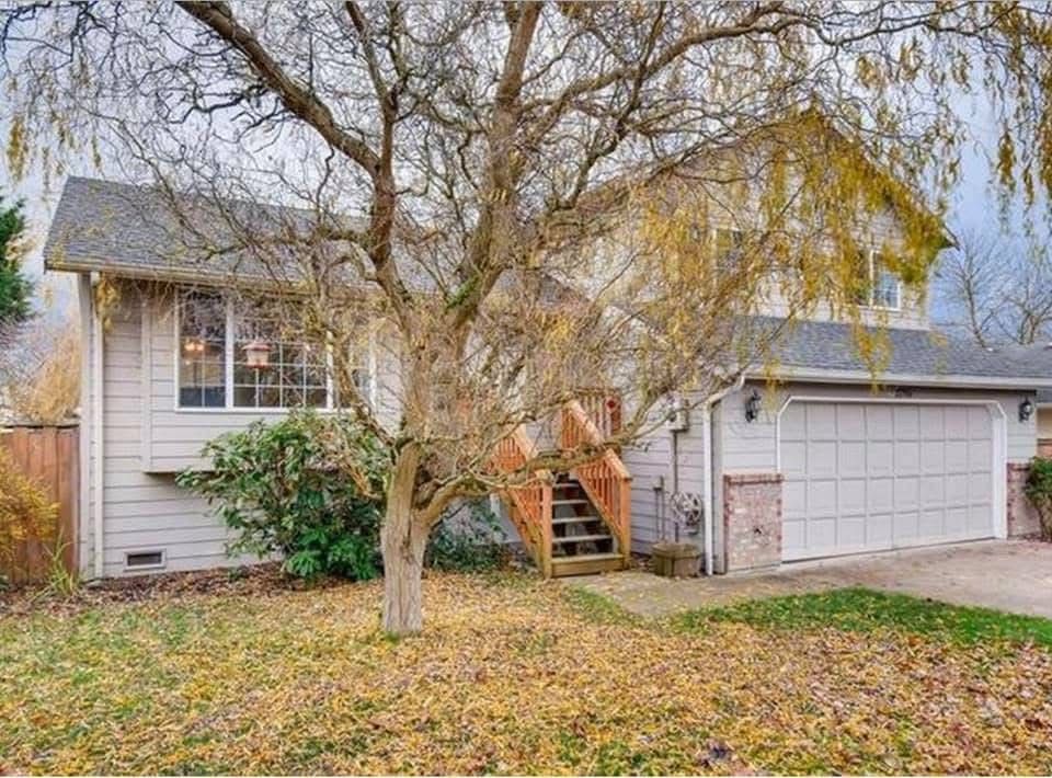 Two-story beige house with a garage, large tree in front, and autumn leaves in the yard