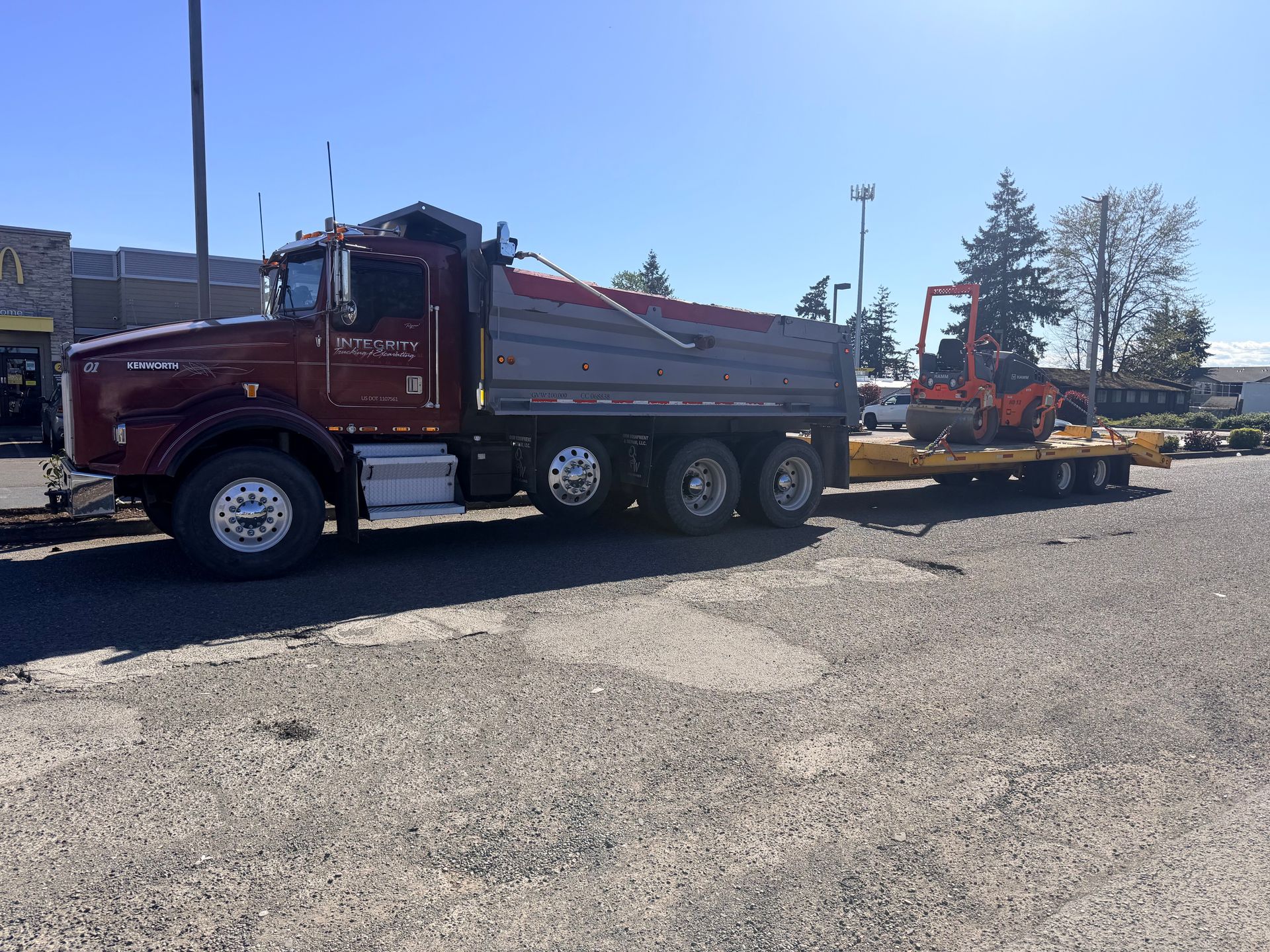 Maroon dump truck towing red construction equipment in a parking lot under a clear blue sky