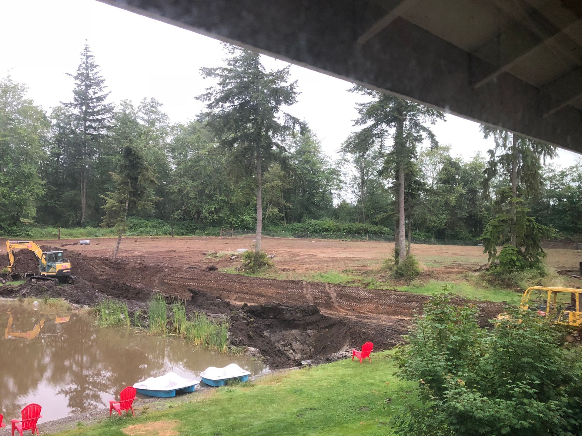 Excavation site with muddy water, dirt embankments, construction equipment, and trees under a bridge.