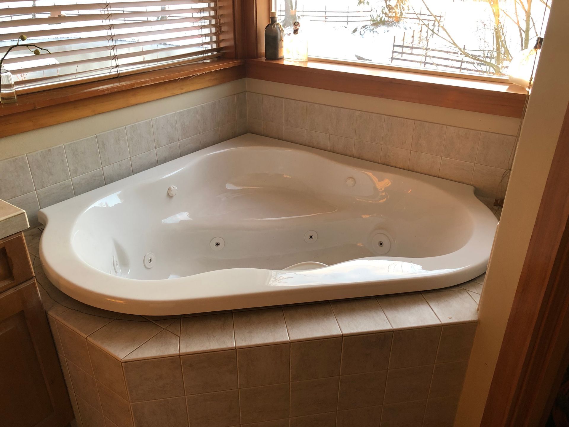 Corner bathtub in a tiled bathroom beneath a window with wooden blinds.