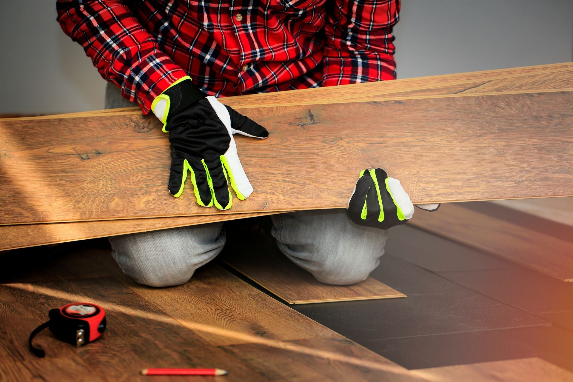 Person in red plaid shirt installing a wooden plank floor with black gloves and a tape measure nearby