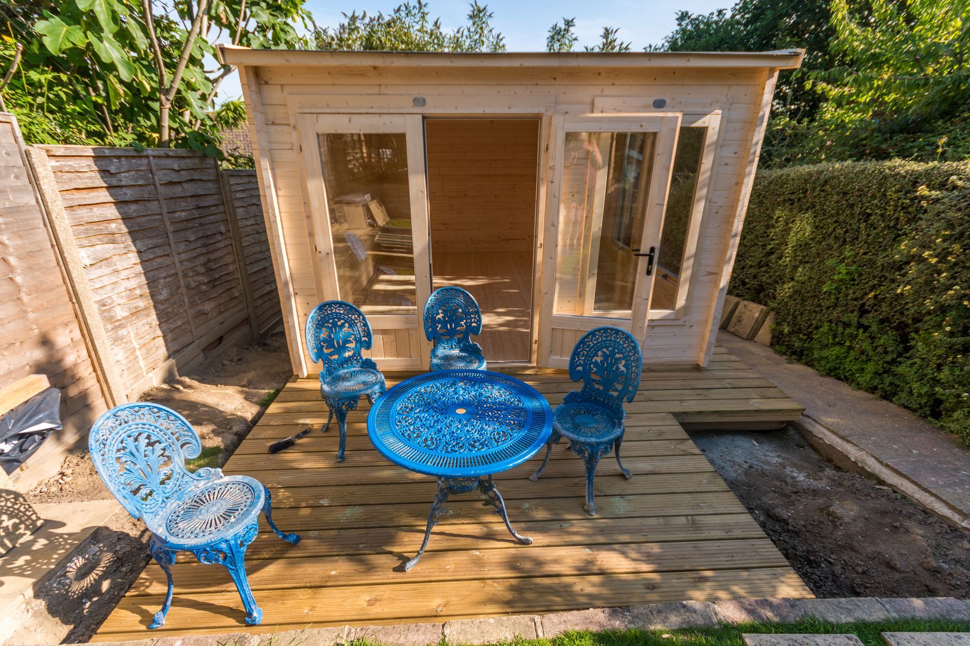 Blue patio table and chairs on a wooden deck outside a small glass garden shed