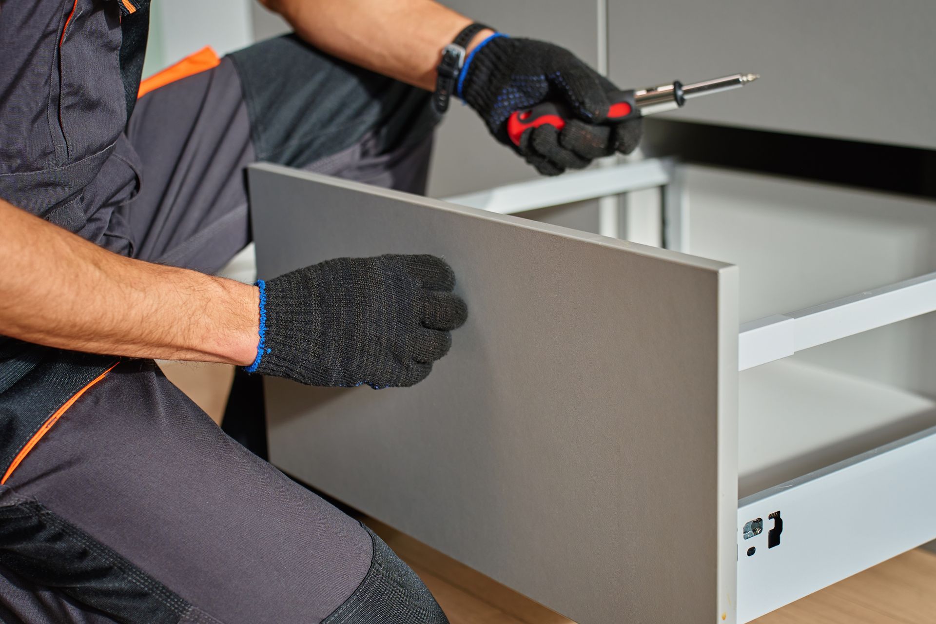 Person assembling a gray drawer in a cabinet, using a screwdriver and gloved hands