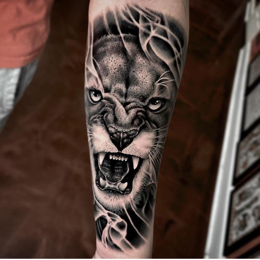 Black-and-gray roaring lion tattoo on a forearm, with detailed smoke-like linework.