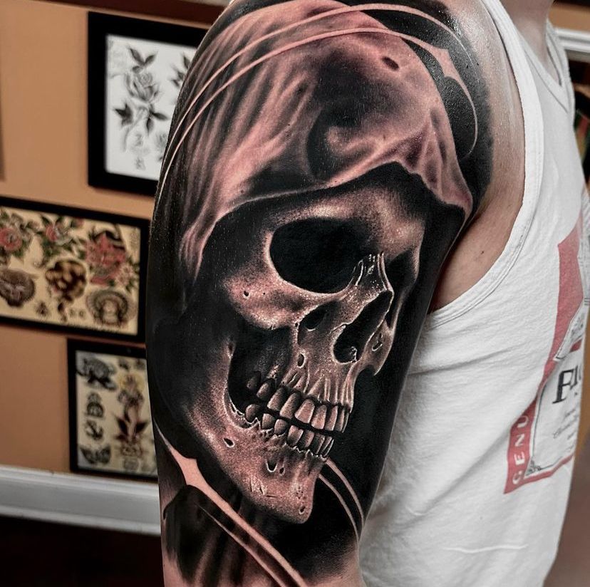 Close-up of a black-and-gray skull tattoo on an upper arm, with framed art on the wall behind it