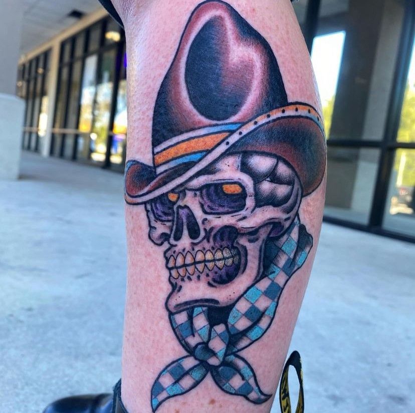 Black-and-gray cowboy skull tattoo with a tilted hat and blue checkered scarf on a forearm