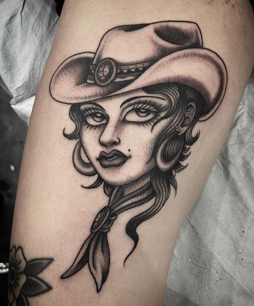 Black-and-gray tattoo of a woman in a cowboy hat with a scarf on a leg
