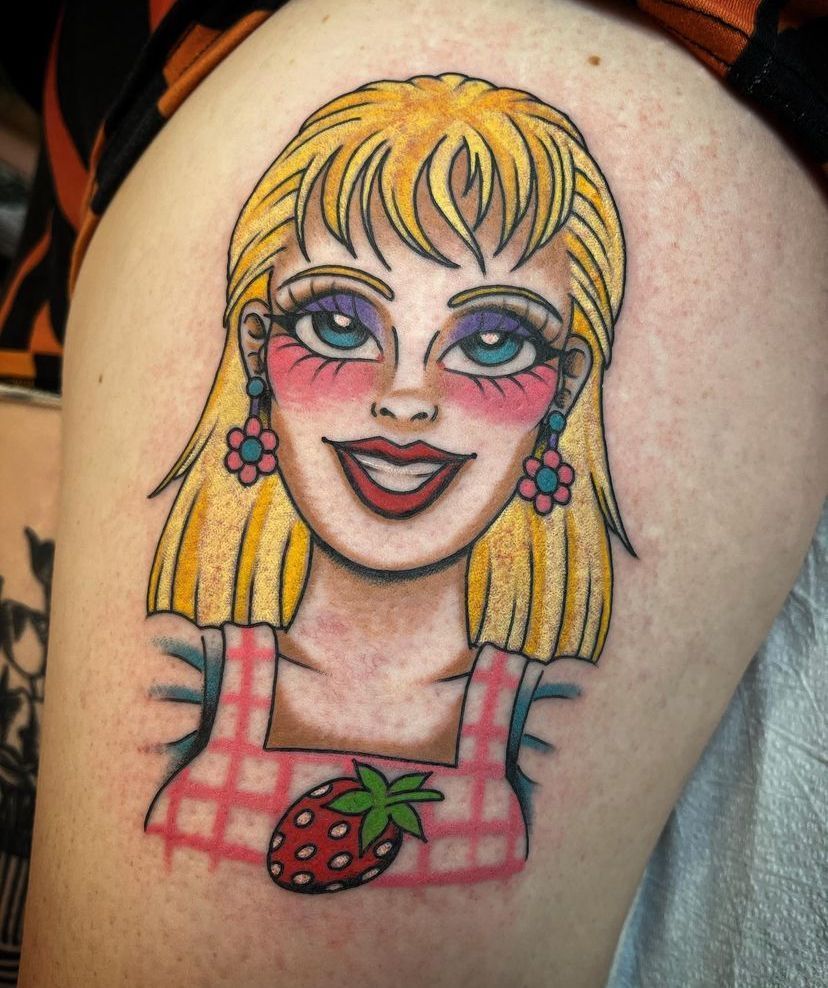Colorful cartoon-style tattoo of a smiling blonde woman in a pink plaid dress with a strawberry emblem.