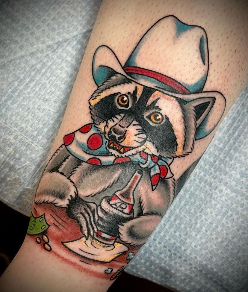 Tattoo of a raccoon in a cowboy hat and bandana, sitting with a red cup on a forearm.
