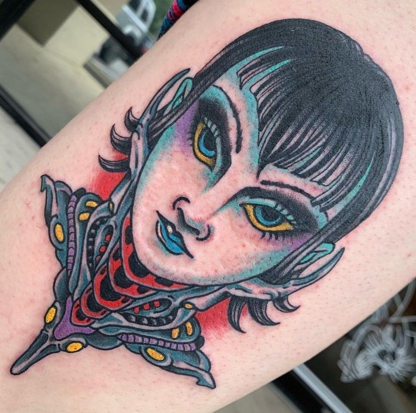 Colorful tattoo of a stylized face with black hair, yellow eyes, and ornate neck details on skin