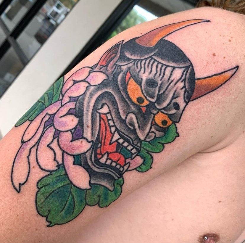 Close-up of a colorful oni mask tattoo with orange horns, pink flowers, and green leaves on skin