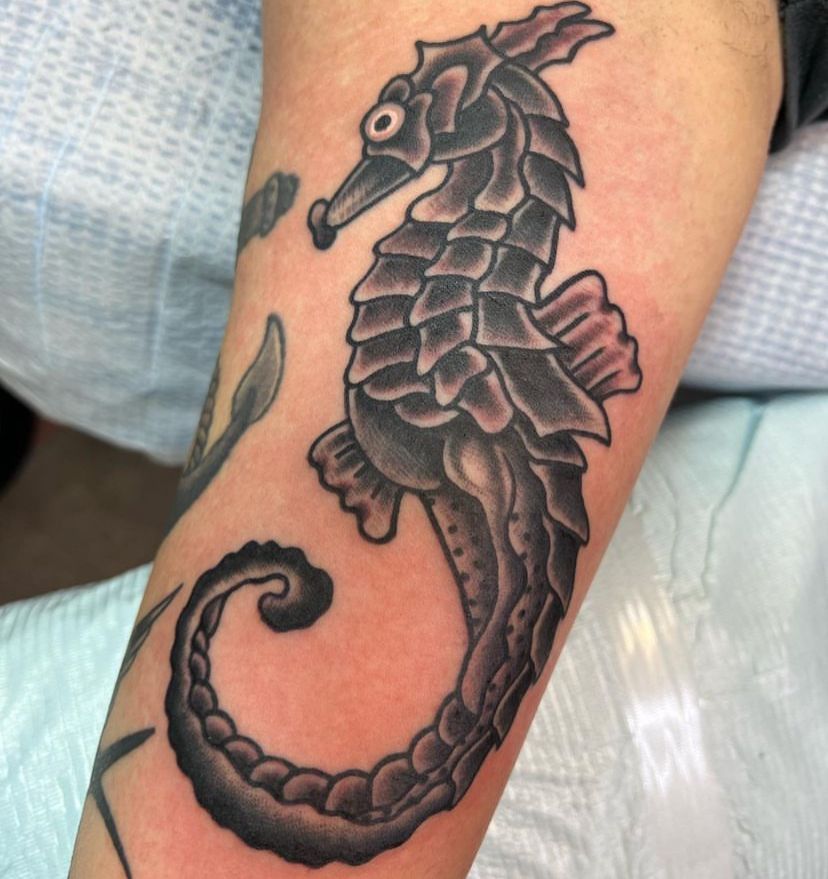 Black seahorse tattoo with a curled tail on a person's forearm