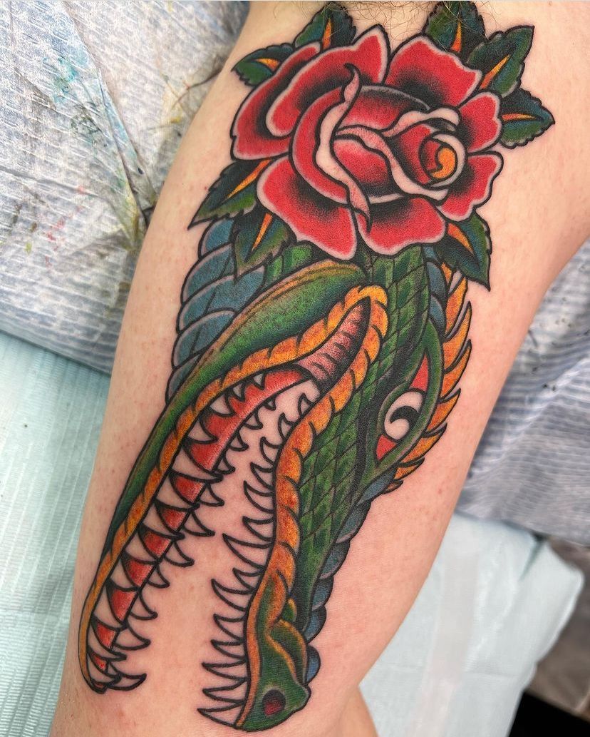 Colorful tattoo of a green crocodile with open jaws and a red rose on an arm