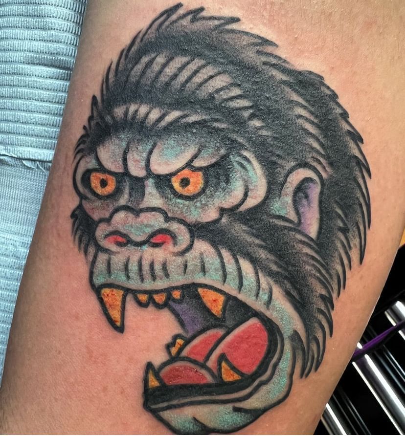 Colorful tattoo of a snarling gorilla face with orange eyes, open mouth, and visible teeth