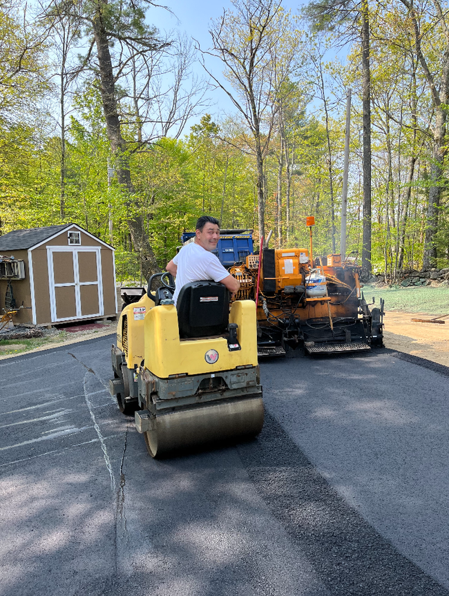 Man on yellow compact roller paving a driveway beside an orange asphalt machine and a shed in a wooded yard