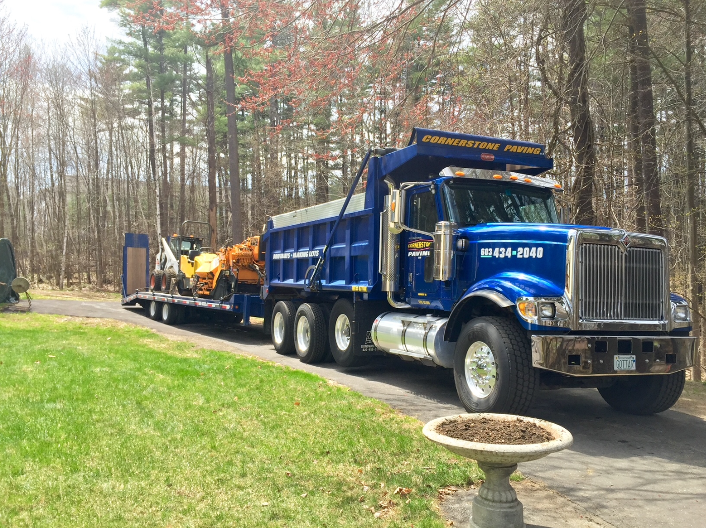 Blue dump truck hauling equipment on a driveway beside a grassy yard and trees