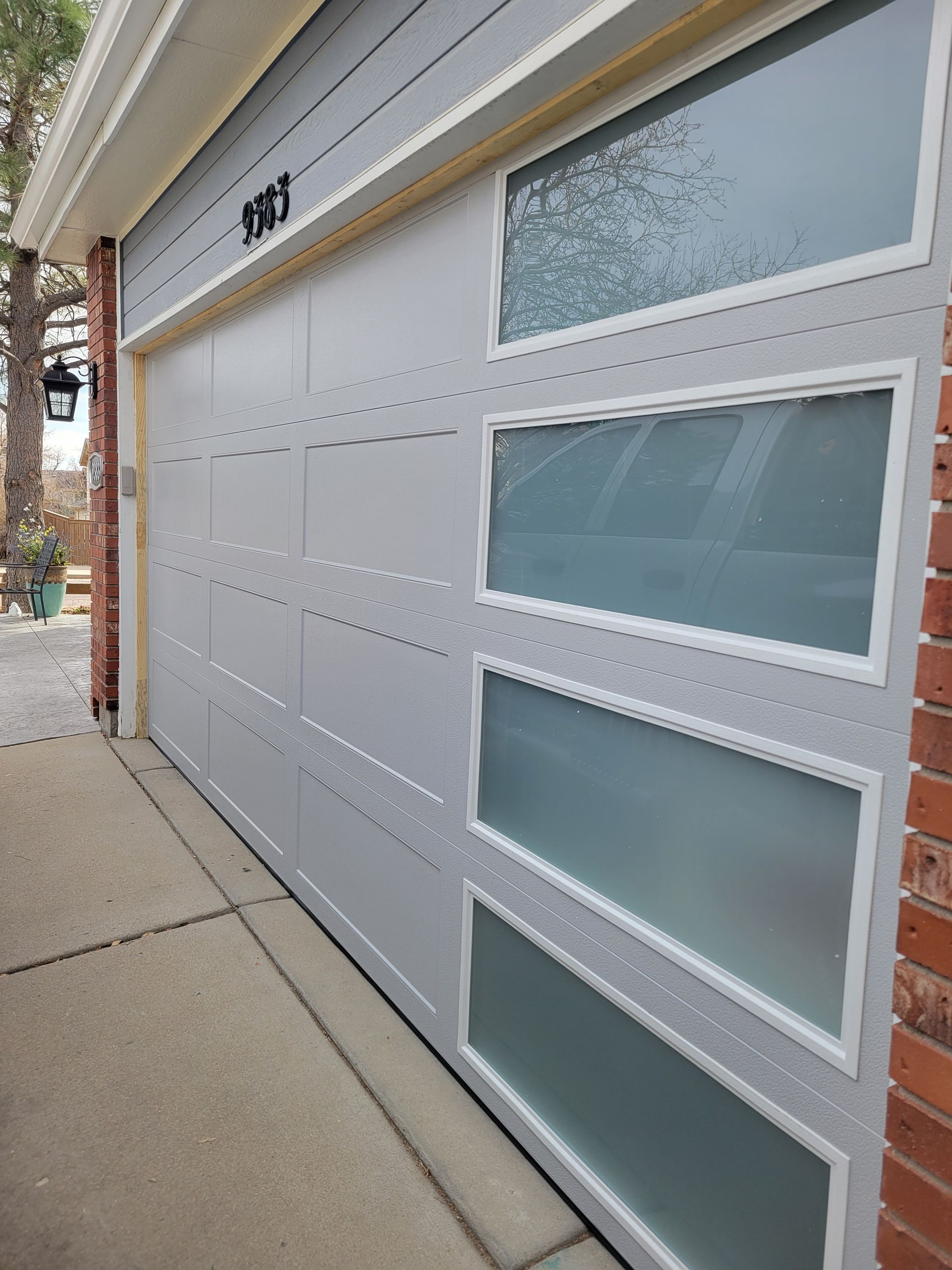 Modern grey garage door with a vertical row of four frosted glass windows on a home with brick siding.