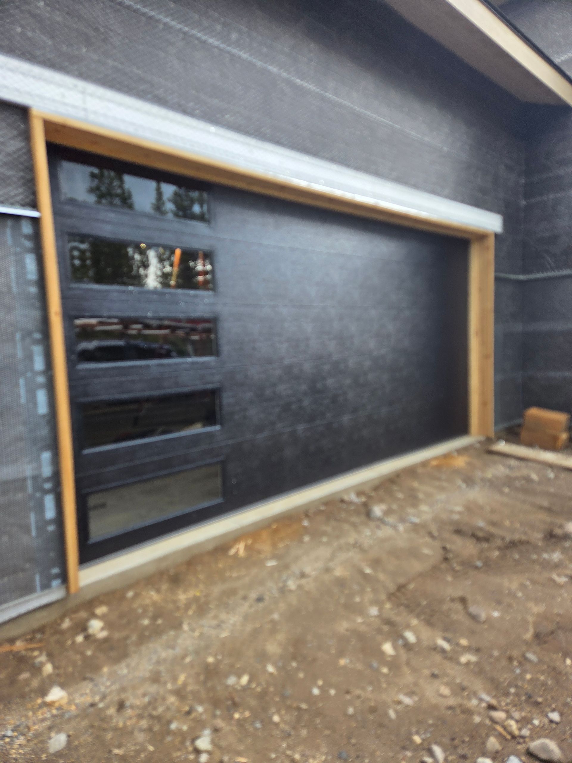 A modern, dark-finished garage door with a vertical row of rectangular glass windows, set in a wood-framed opening.