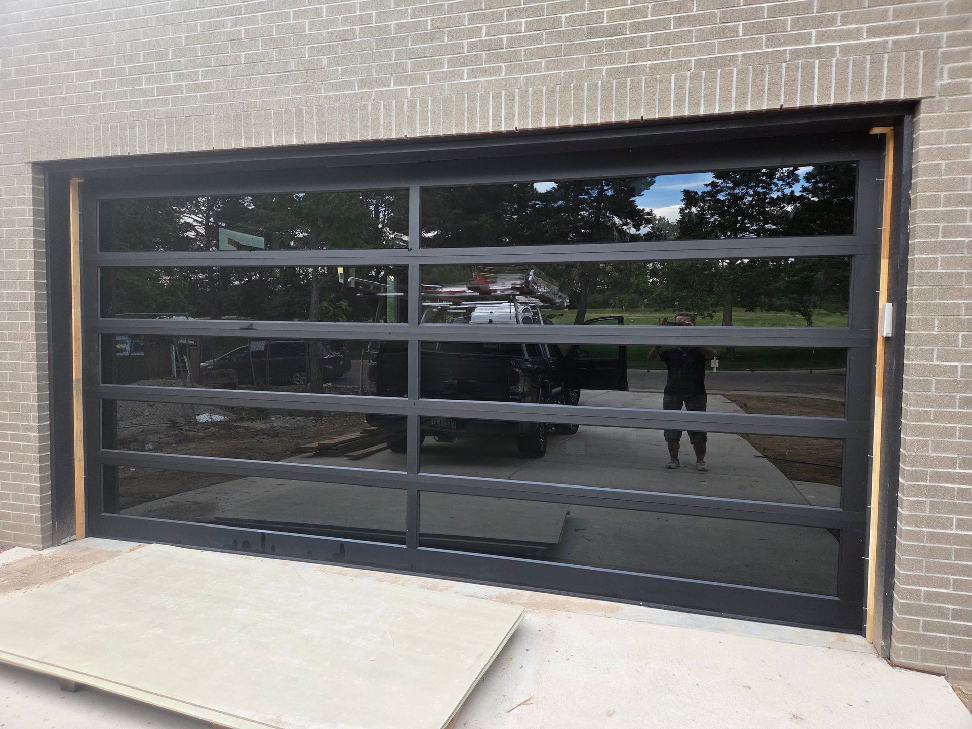 A modern black-framed glass garage door installed in a light brick exterior wall, reflecting a driveway and trees.