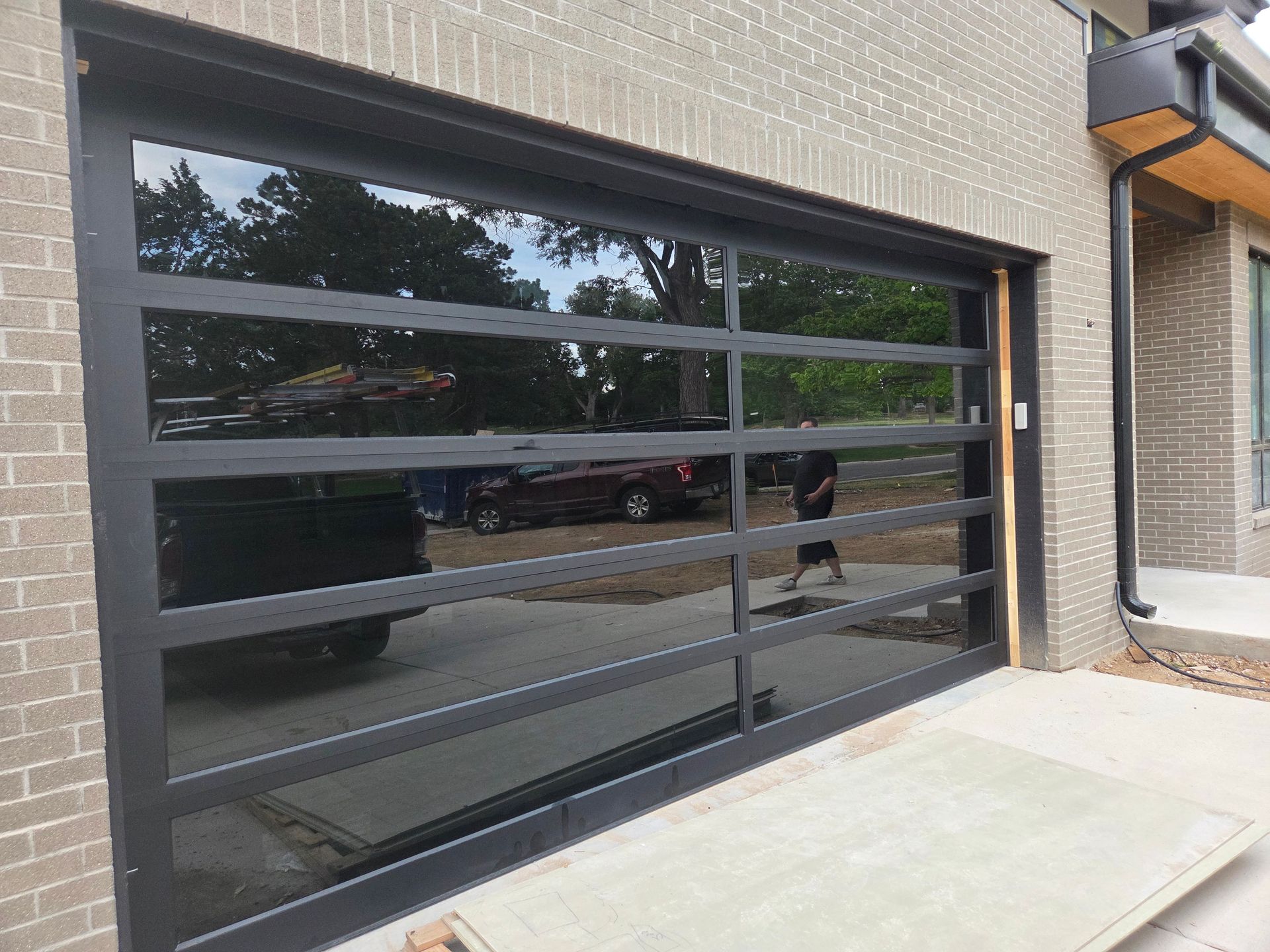 A modern, black-framed garage door with horizontal glass panels reflecting a driveway and trees outside a brick house.