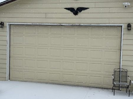 A beige garage door featuring a black eagle wall decoration, with snow on the ground and a metal chair to the right.