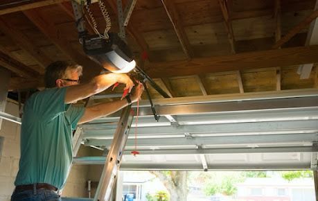 A person standing on a ladder in a garage, repairing a garage door opener motor mounted to the ceiling.