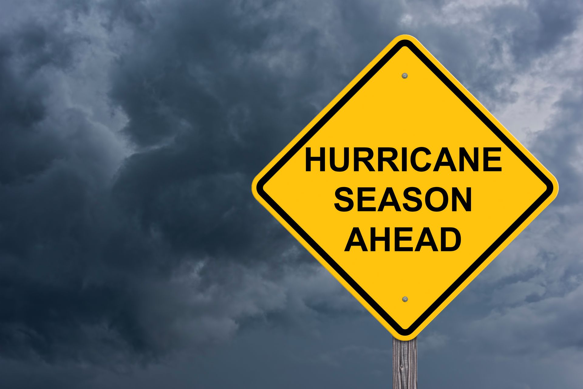 A yellow warning sign that reads, “Hurricane Season Ahead”, with a stormy sky background