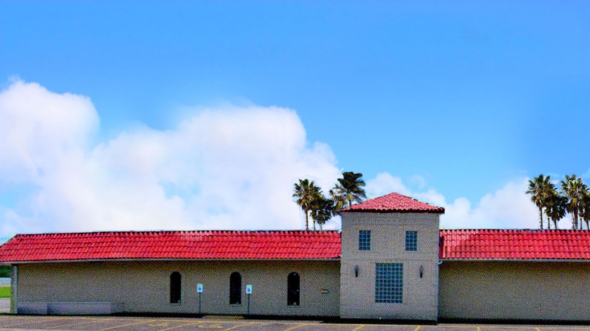 Building with red tile roof, tower, and palm trees against a blue sky with clouds.