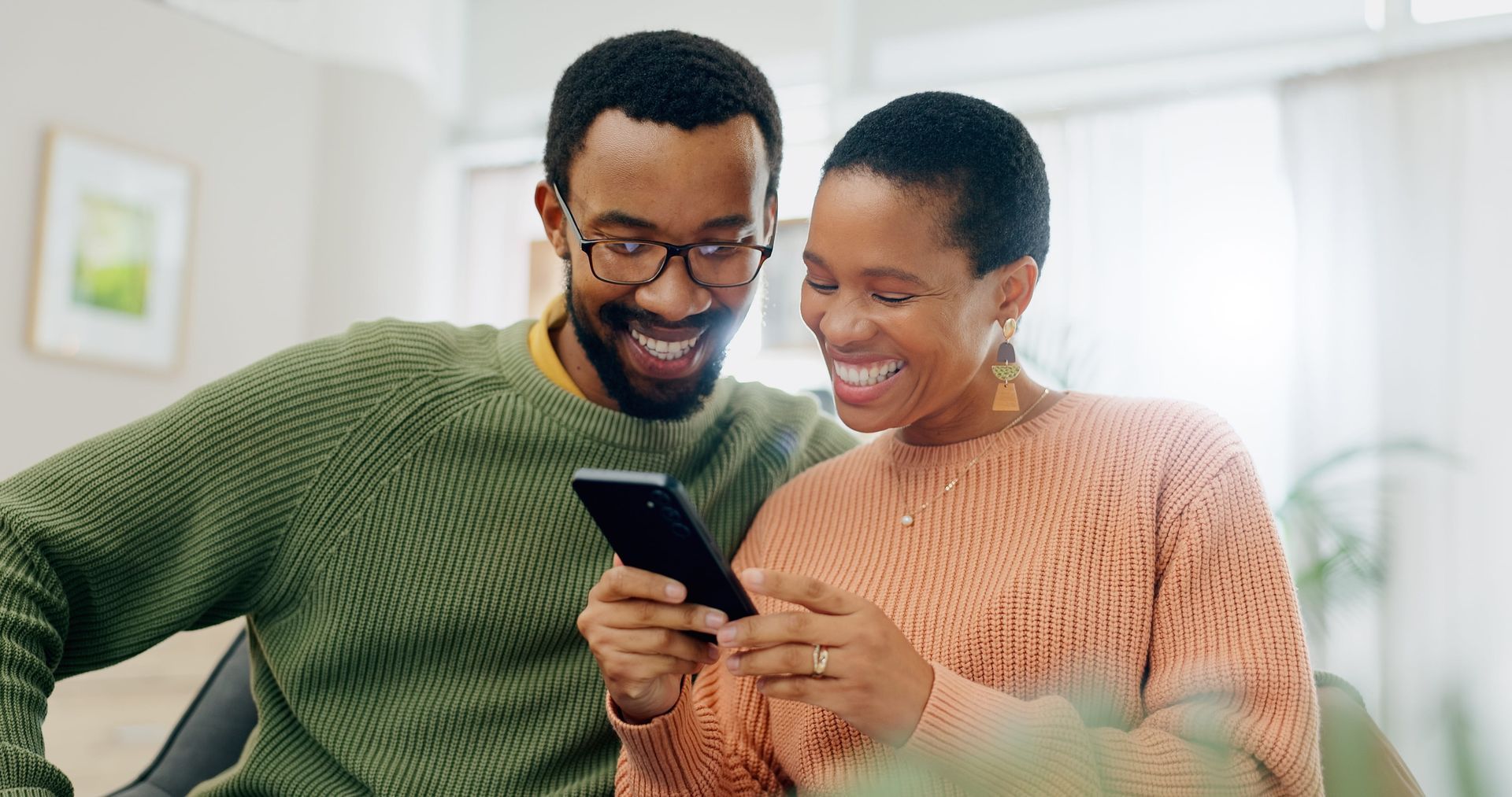 Two people smiling and looking at a smartphone together on a couch indoors