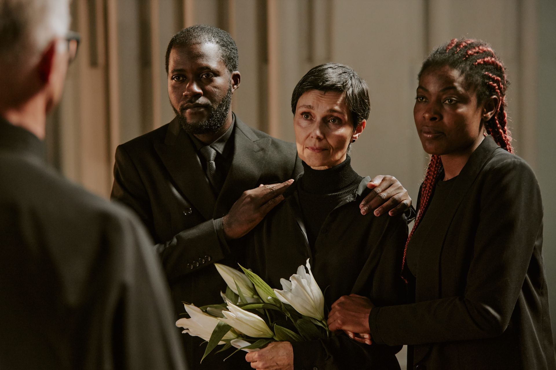 Three mourners in black stand together holding white lilies at a funeral