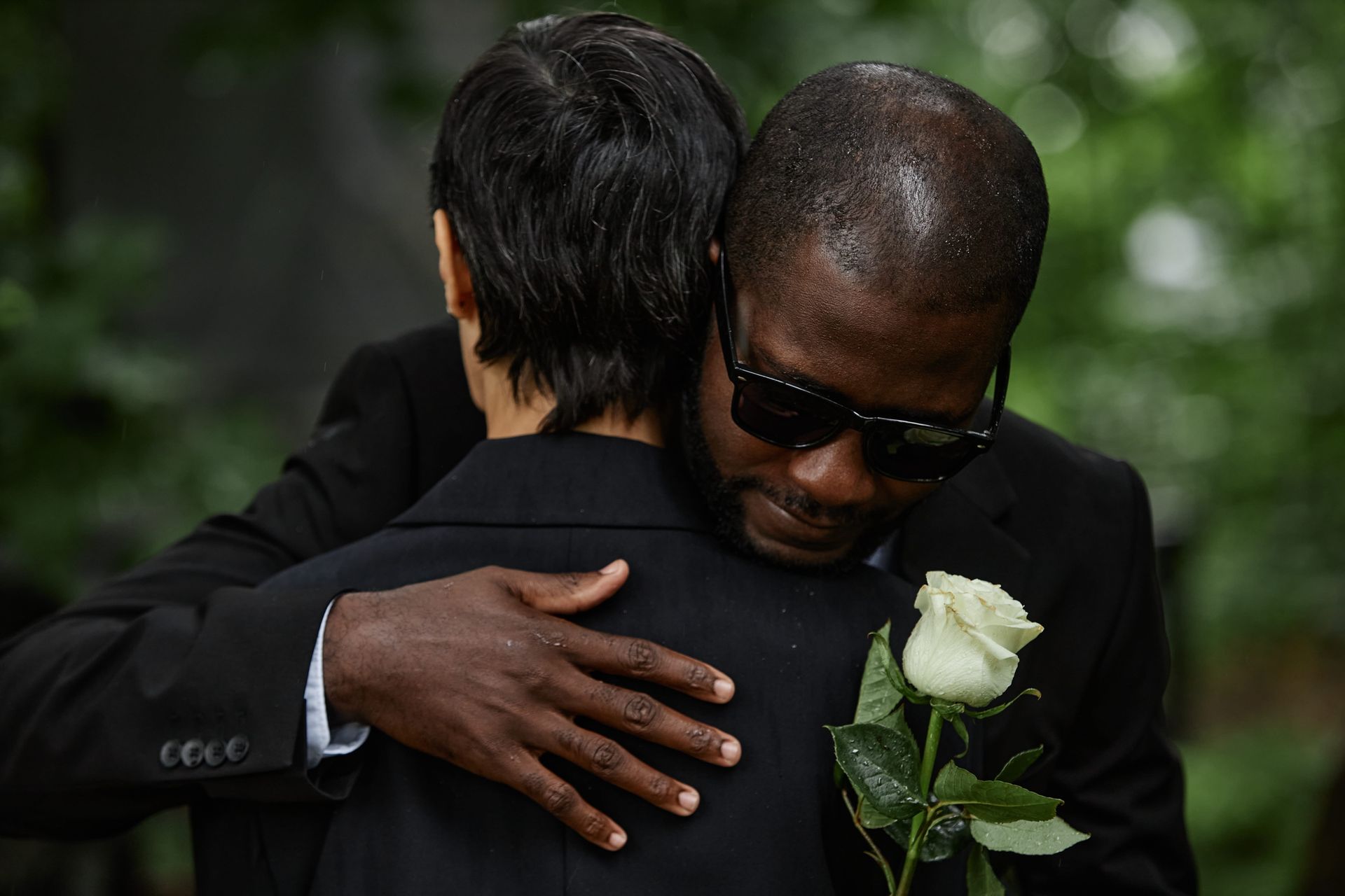 Two people embracing in black formal wear, one holding a white rose, in a somber outdoor setting
