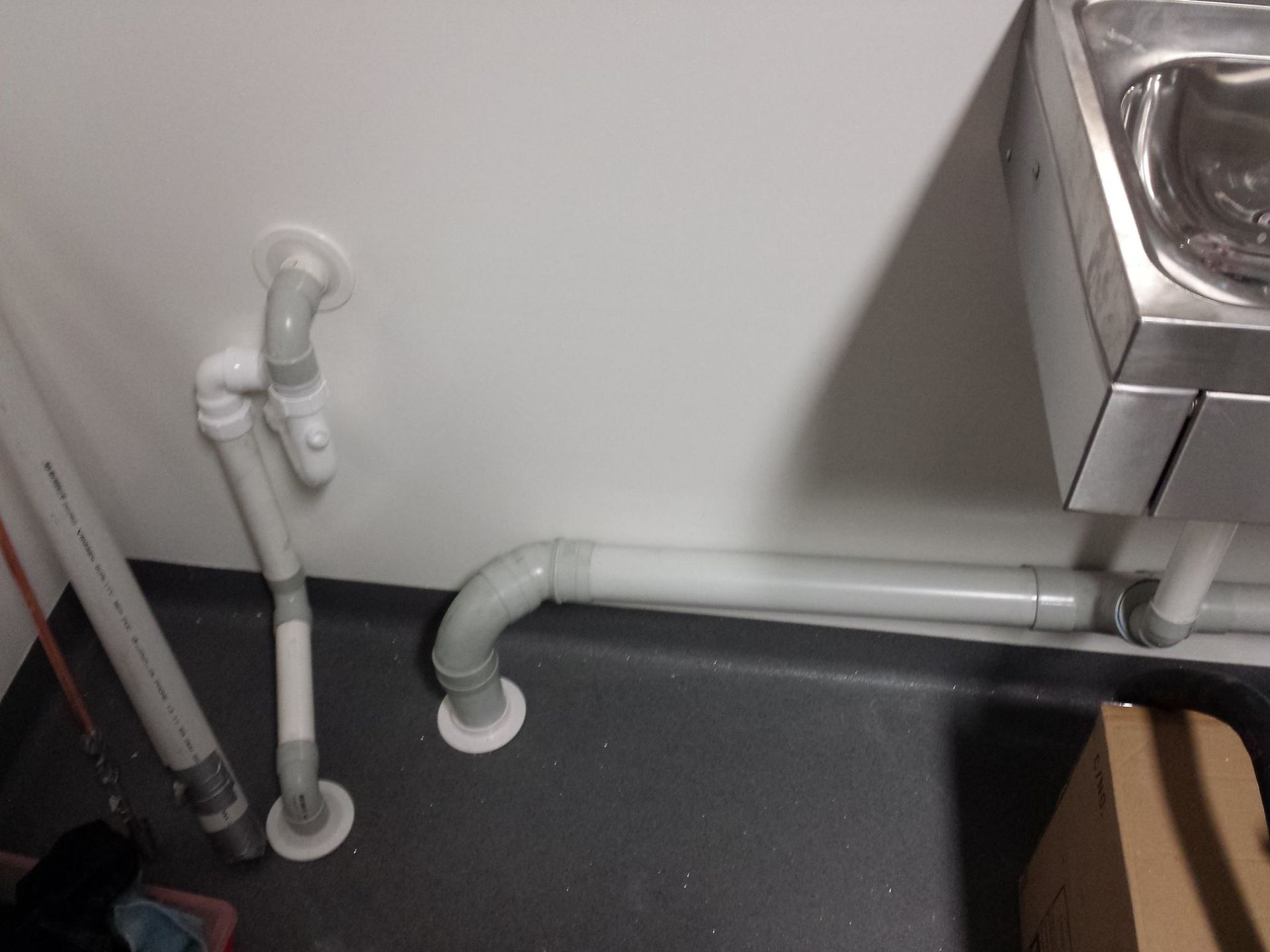 Stainless steel triple sink in commercial kitchen. White plumbing pipes beneath sinks. Blue broom leans against the sink.