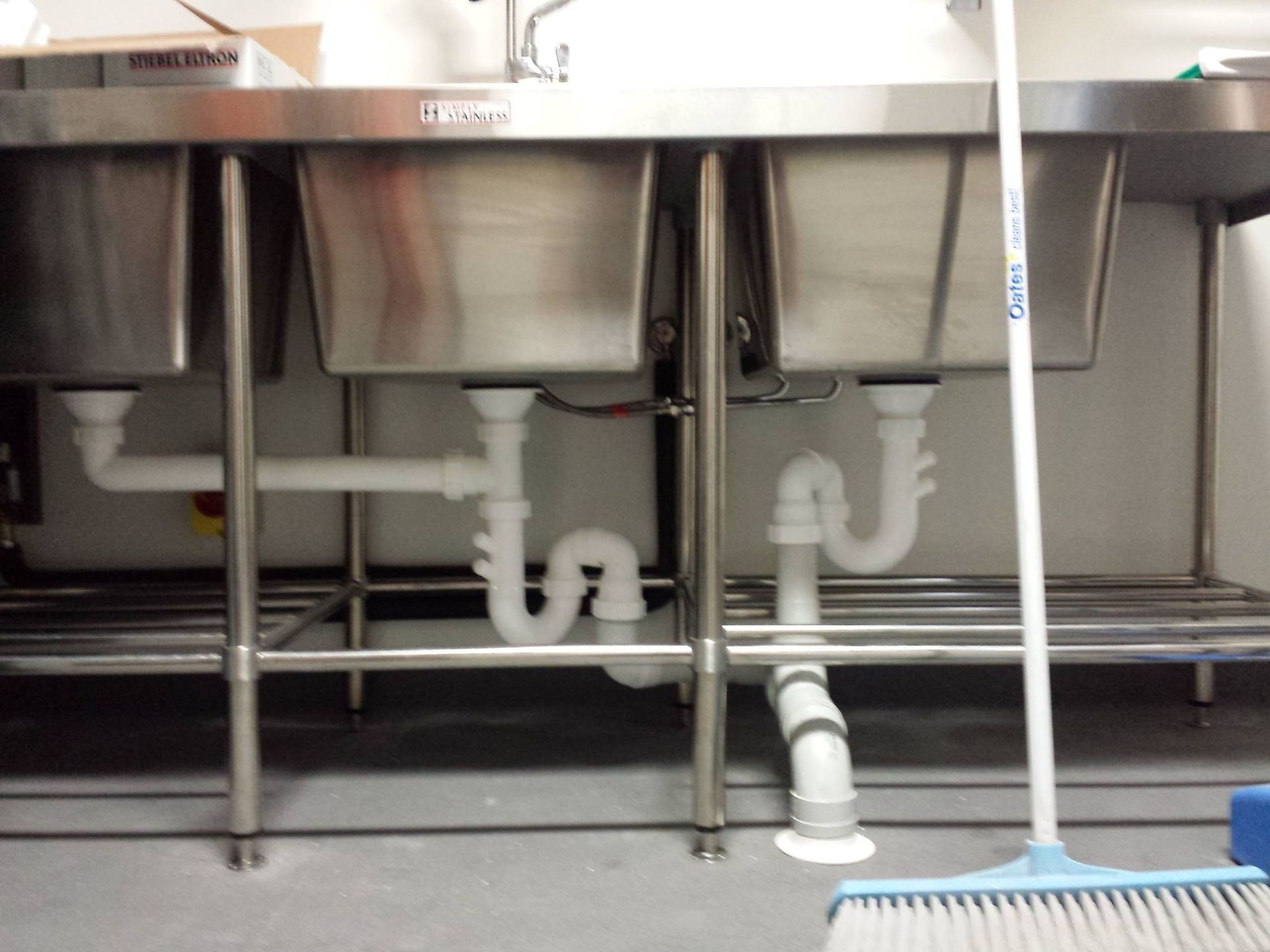 Stainless steel triple sink in a commercial kitchen with plumbing visible. A broom is leaning against the right side.
