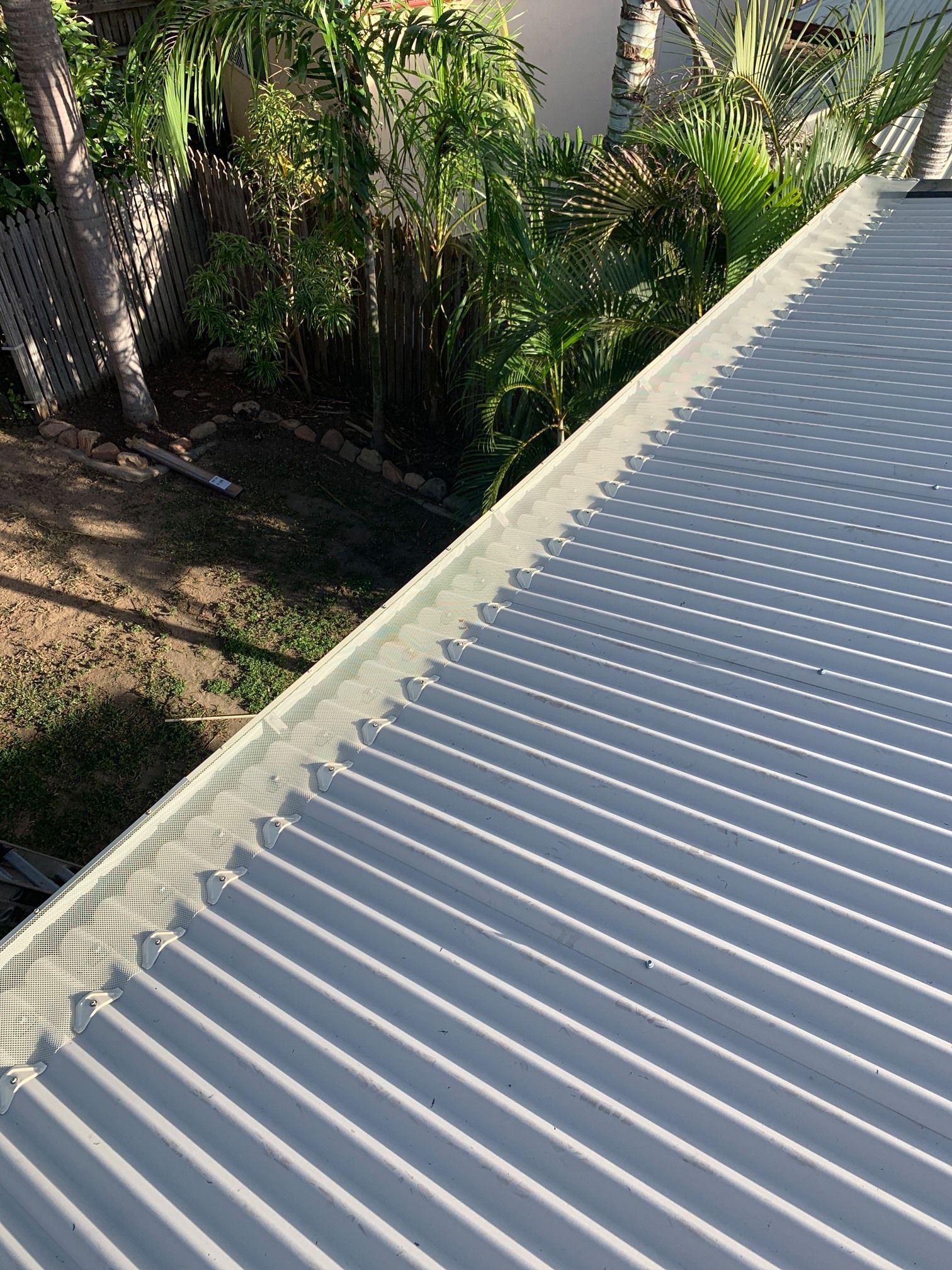 A corrugated metal roof edge in sunlight, with trees and a yard in the background.