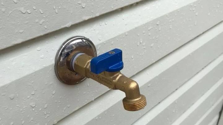 Brass outdoor water faucet with blue handle on white siding.