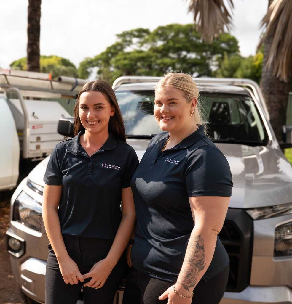Two women in work shirts stand by a silver truck outdoors.