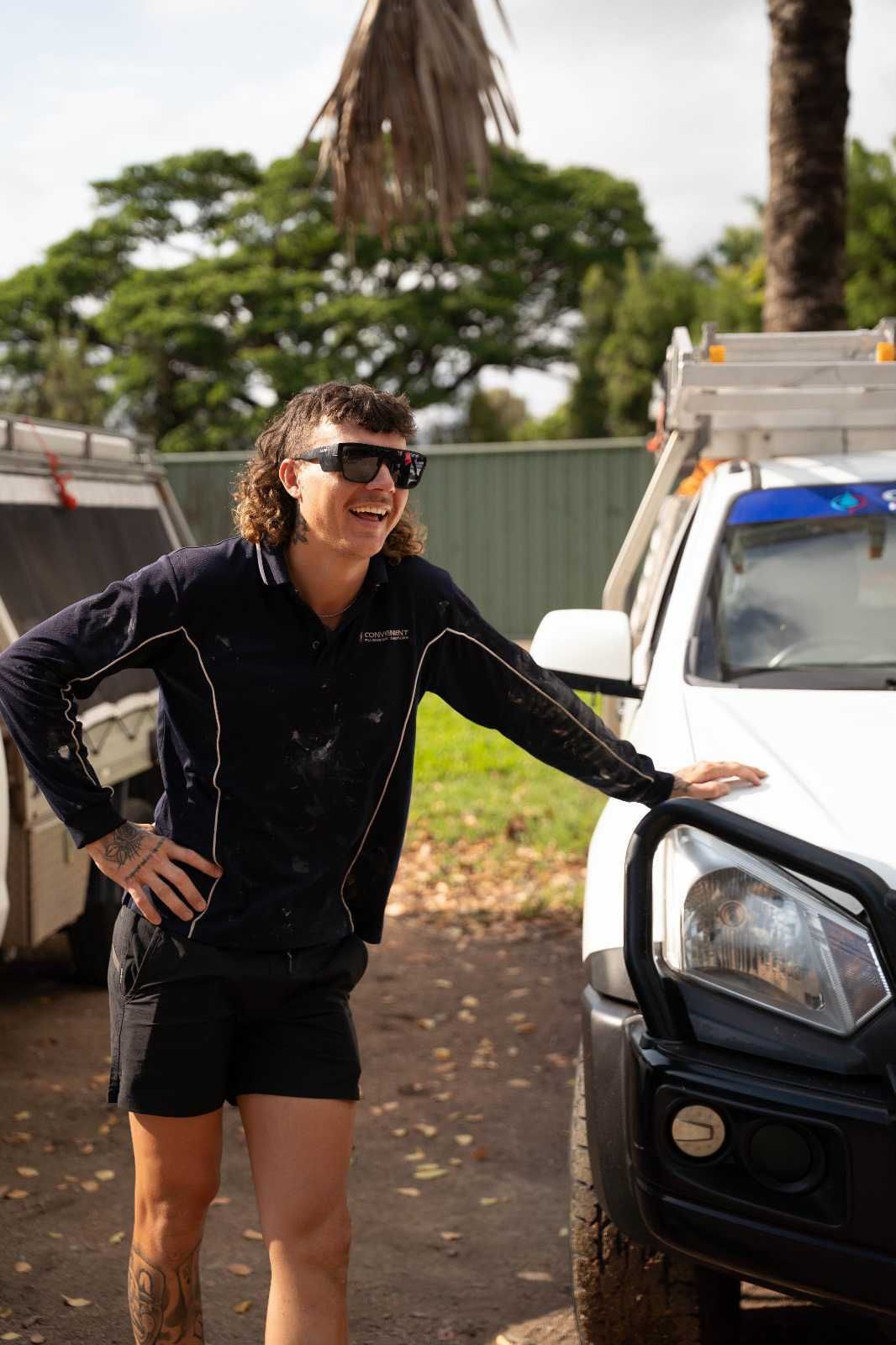 Team member with a mullet, sunglasses, and work clothes smiles, leaning on a white truck. Outside.