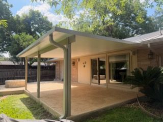 Covered backyard patio with beige siding, green posts, and a sliding glass door under trees.
