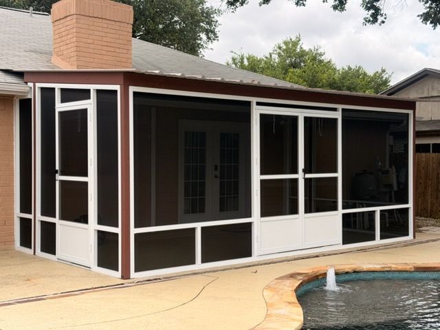 Backyard sunroom with white-framed windows on a brick house, beside a grassy yard and black storage bin.