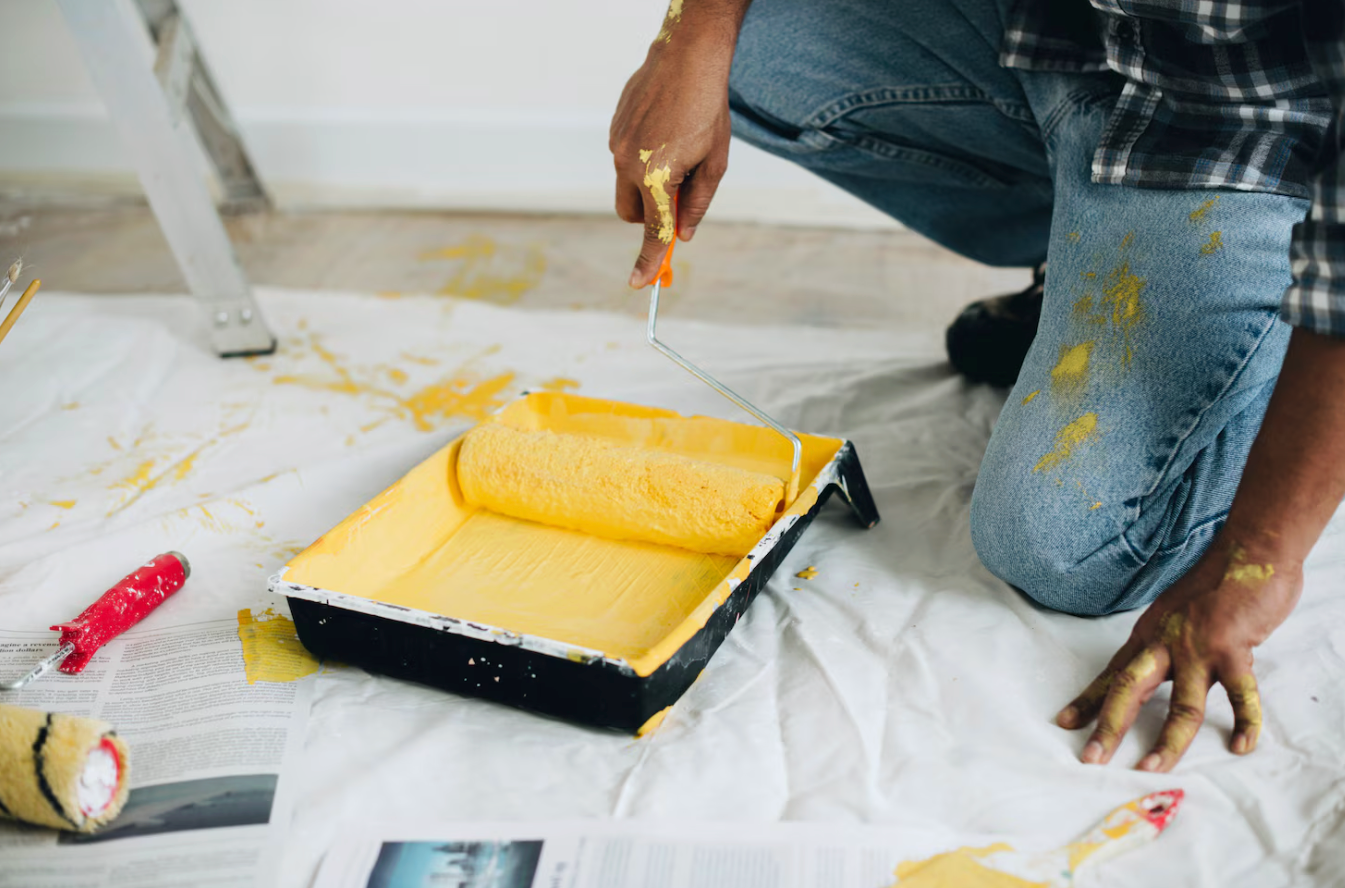 Person painting a wall with yellow paint using a roller, with paint tray and drop cloth on the floor