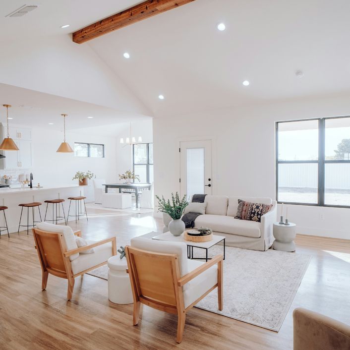Bright minimalist living room with white sofa, tan chairs, rug, and open kitchen with pendant lights