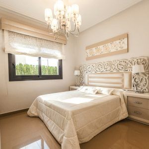 Bright bedroom with a white ornate bed, chandelier, window, and beige walls and floor.
