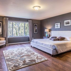 Modern bedroom with gray walls, a large window, bed, nightstand, lamp, and floral area rug