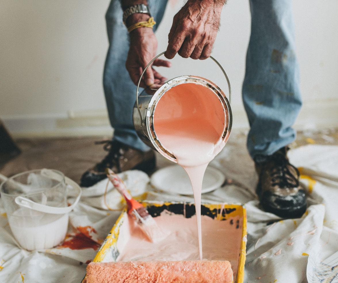 Person pouring pink paint from a bucket onto a drop cloth while painting a floor indoors