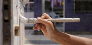 Hand holding a paintbrush applying white paint to a metal railing outdoors