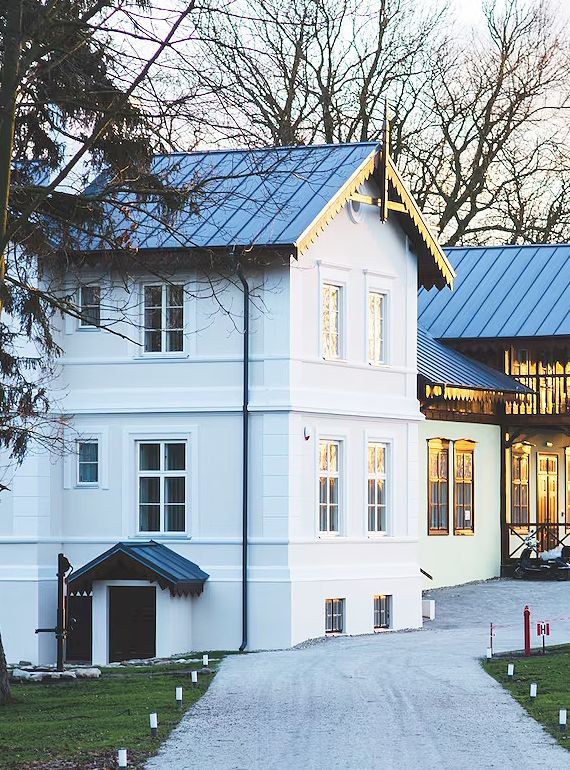 White two-story house with a blue roof and lit windows along a paved driveway at dusk