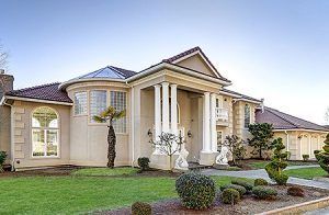 Large beige suburban house with columns, a tiled roof, and a manicured front lawn under a clear sky